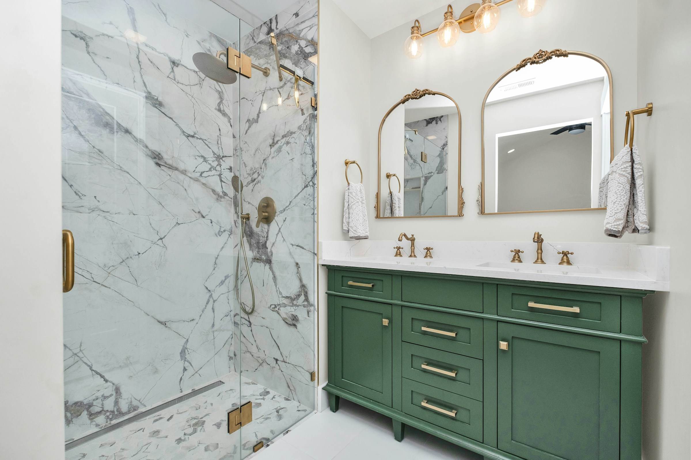 Green double vanity with brass fixtures beside a marble-clad glass shower