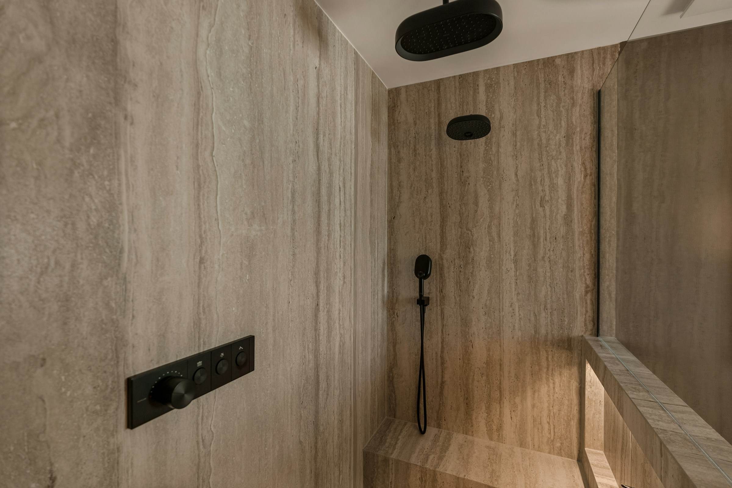 Travertine-clad walk-in shower with a matte black thermostatic control panel and bench